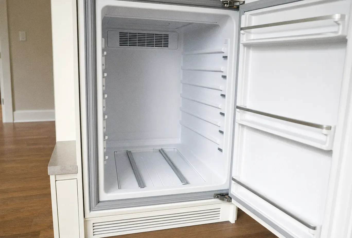 An empty white undercounter refrigerator compartment with the door open, showing bare shelf rails and a vent grille at the back